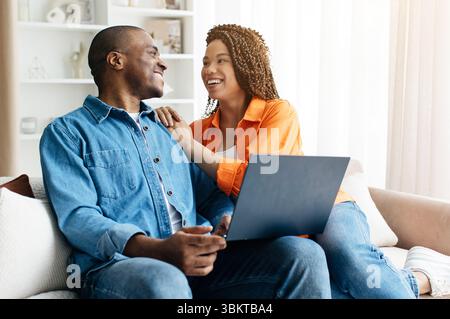 Black couple smiling at each other while sitting on couch with laptop Stock Photo
