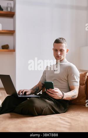 Vertical shot of focused Caucasian young adult man sitting on couch using laptop and smartphone multitasking for business purposes while working at home office Stock Photo