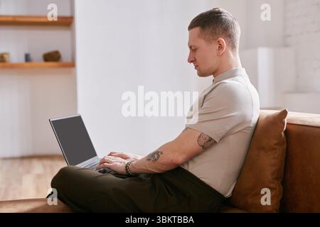 Side view of focused Caucasian young adult man sitting on sofa typing on laptop while working at home office Stock Photo