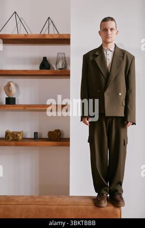 Vertical shot of young adult Caucasian man standing on sofa in business setting wearing oversized suit jacket and trousers, looking directly at camera with neutral expression Stock Photo