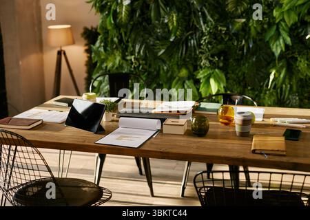 Wooden table in modern office setting displaying digital tablet, notebooks, documents, reusable coffee cup, and stationery with green plant wall background Stock Photo