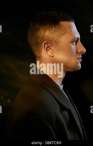 Vertical shot of Caucasian young adult man standing in profile view against dark background, showing short hair and clean facial features in studio Stock Photo