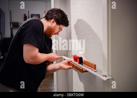 Focused man aligns a wooden bracket on a wall using a spirit level and pencil, working on a DIY home improvement task. Stock Photo