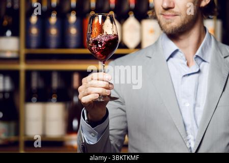 Vintner man tasting still red dry wine from a glass in cellar. Close up shot of male sommelier hand that hold the wine glass on shelf background Stock Photo