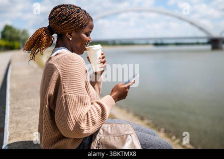 Young black woman is using smartphone and drinking coffee to go, outdoor by the river. Stock Photo