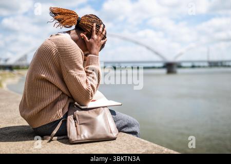 Portrait of displeased young black woman who is sitting by the river in the city. Stock Photo