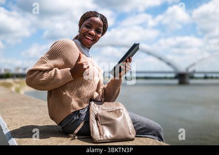 Young black woman is relaxing outdoor by the river. She is using digital tablet and showing thumbs up. Stock Photo