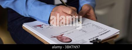 Middle aged Caucasian woman filling out criminal profile form on clipboard, holding pen and writing details, close up of hands and paperwork, prisoned context indicated by document Stock Photo