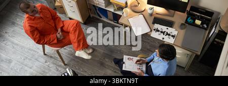 Caucasian young adult woman wearing orange prison uniform sitting with hands folded meeting middle aged Caucasian woman psychologist taking notes during prison counseling session Stock Photo