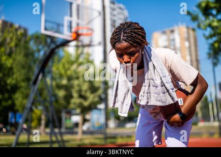 Young black woman is standing on basketball court. She is tired of playing and taking a break. Stock Photo