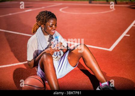 Young black woman practicing basketball alone at sports court in the morning. She is holding bottle of water and taking a break. Stock Photo