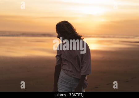 Silhouette of woman on a beach at sunset, looking down thoughtfully Stock Photo