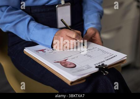 Middle aged Caucasian woman filling out criminal profile form on clipboard, holding pen in right hand, wearing identification badge, sitting indoors, close up on hands and document Stock Photo