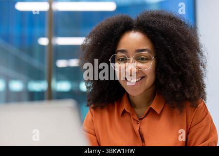 Close-up of a smiling woman with glasses working on a laptop in a modern office setting Stock Photo