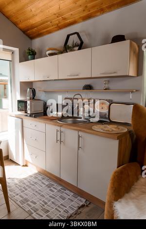 A bright, inviting small kitchen features light beige cabinets with wooden countertops, a stainless steel sink, microwave, toaster, and kettle, evokin Stock Photo