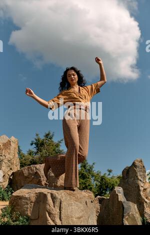 Curly-haired young woman showcases her summer style while playfully posing on rocks outdoors. Stock Photo