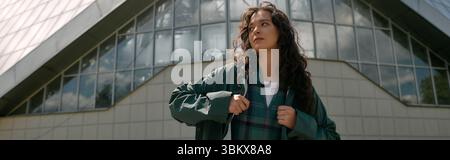 Curly-haired woman showcases trendy fashion in stylish raincoat outdoors under a sunny sky, banner Stock Photo