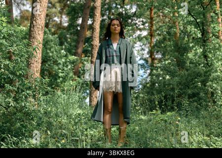 Curly-haired woman showcases trendy outfit while posing amidst lush greenery on a summer day. Stock Photo