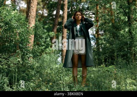 Curly-haired woman showcases her fashion sense in a stylish raincoat while outdoors in the woods. Stock Photo