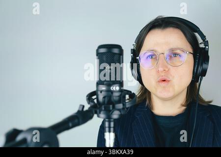 Young caucasian female singing into microphone wearing headphones and glasses. Stock Photo