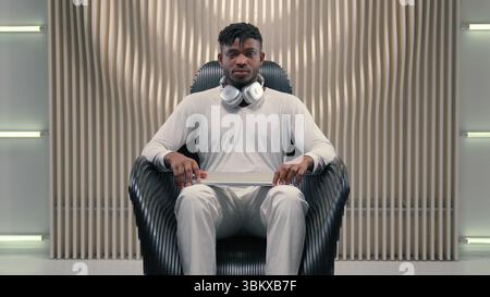 Young African American man guy male put on headphones opening laptop indoors in futuristic room looking at screen concentrated in modern cyberspace Stock Photo