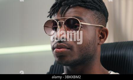 Portrait African American guy man ethnic male putting on sunglasses glasses wearing looking at camera sitting in futuristic white studio futurism Stock Photo