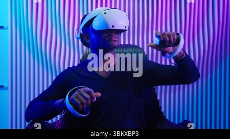 Cyber gamer African American man in VR glasses play virtual reality game in neon futuristic space indoors dark blue room guy male cyberpunk fashion Stock Photo