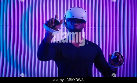 Smiling happy African American man in VR glasses play virtual reality game in neon futuristic space browsing interactive future software interface guy Stock Photo