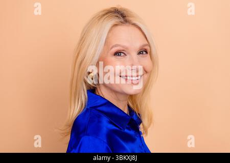 Confident mature businesswoman wearing a stylish blue shirt, smiling warmly against a soft beige background, showcasing elegance Stock Photo