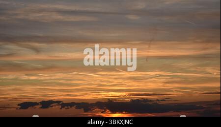 Warm light of the setting sun illuminates scattered clouds with vibrant golden, orange, blue, and soft gray hues Stock Photo