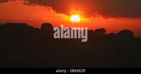 Fiery sunset sky with warm orange, gray, and yellow hues above town silhouettes and soft clouds drifting in twilight. Stock Photo