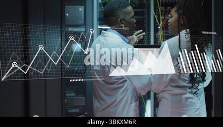 Gesturing engineers in lab coats reviewing metrics on laptop in data center, with racks and cables Stock Photo