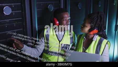 Pointing technicians explaining code overlay at data center racks, with laptop and red headsets Stock Photo