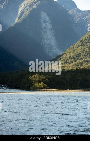 A forested shoreline catches golden light at the base of towering, shadow-draped mountains, where steep rocky faces rise above rippling water, creatin Stock Photo