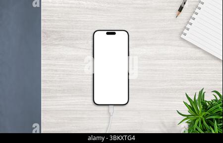 Top view of smartphone on charger with blank screen, placed on desk with notebook and plant. Ideal for productivity and app mockups Stock Photo