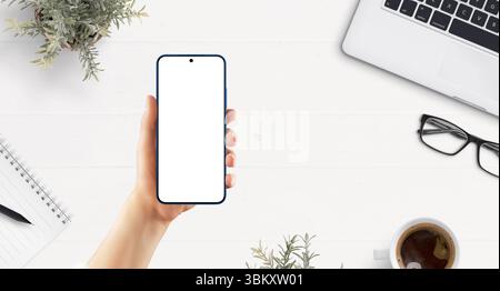 Top view of smartphone in female hand over white desk with laptop, coffee, notebook, and workspace items. Ideal for app mockup or UI display Stock Photo