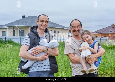 Elderly man holding smiling granddaughter with plush toy, standing next to his adult son holding infant in suburban yard. Generational family moment i Stock Photo