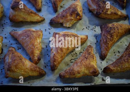 A baking tray filled with freshly baked golden brown triangular cakes. The cakes have a flaky, crunchy texture, are sprinkled with sezam seeds on top. Stock Photo