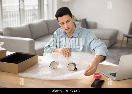 Confident millennial male ecommerce store manager packing cups in parcel Stock Photo