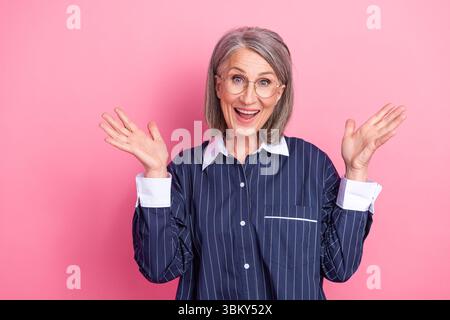 Cheerful senior woman with hands raised looking excited wearing stylish striped shirt against bright pink backdrop Stock Photo