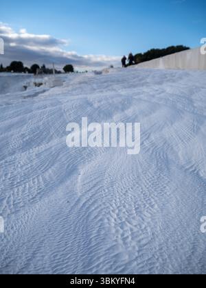A close-up shot of the white, textured surface of Pamukkale, Turkey, with two blurred figures walking in the background, showcasing the unique geological formation and tourist activity. Stock Photo