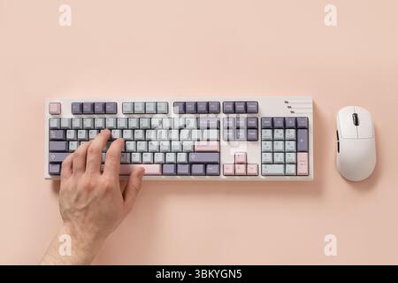Mans hand texting and typing on modern mechanical keyboard and mouse on pink background. Colorful pastel pink and green key cap. View from above. Flat Stock Photo