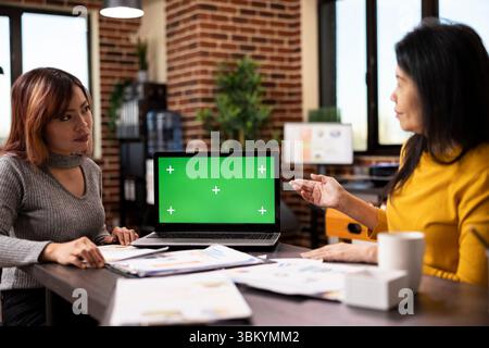 Digital device displaying a blank chromakey template sits on the table alongside scattered paperwork, with two women having a business discussion. Strategy meeting between coworkers. Stock Photo