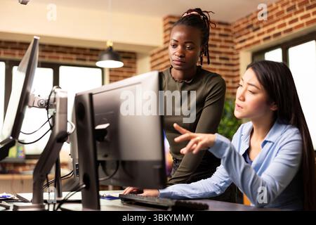 Diverse female designers discussing digital artwork and making creative decisions in modern workspace. Asian and African American colleagues intently reviewing graphic design updates on device screen. Stock Photo