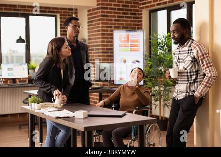 Business professionals engaging in a friendly discussion at office desk, fostering an inclusive and collaborative work environment. Multiethnic coworkers talking and having coffee. Stock Photo