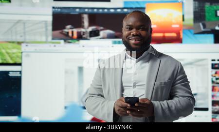 Portrait of cheerful man browsing social media feed on smartphone in front of digital video wall. Happy person using mobile phone, floating icons highlight interactive engagement online, camera A Stock Photo
