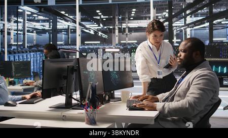 Lead data center admin overseeing IT expert integrating AI in equipment rigs. African american employee under server farm team leader supervision using deep learning for operational tasks, camera A Stock Photo