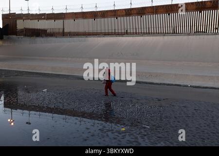 A person in red clothing and a blue backpack walks through shallow water near a tall border fence topped with barbed wire. Ciudad Juarez, Chihuahua. Stock Photo