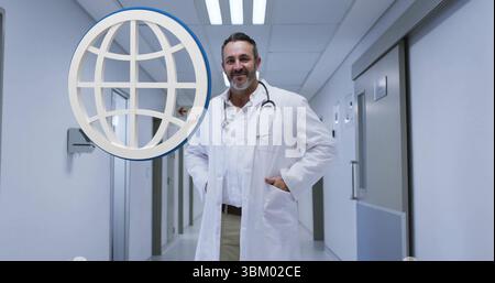 Standing middle-aged doctor wearing lab coat and stethoscope in medical corridor, with globe icon Stock Photo