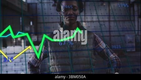 Standing data analyst wearing puffer vest scanning warehouse, overlaid with neon charts and grid Stock Photo